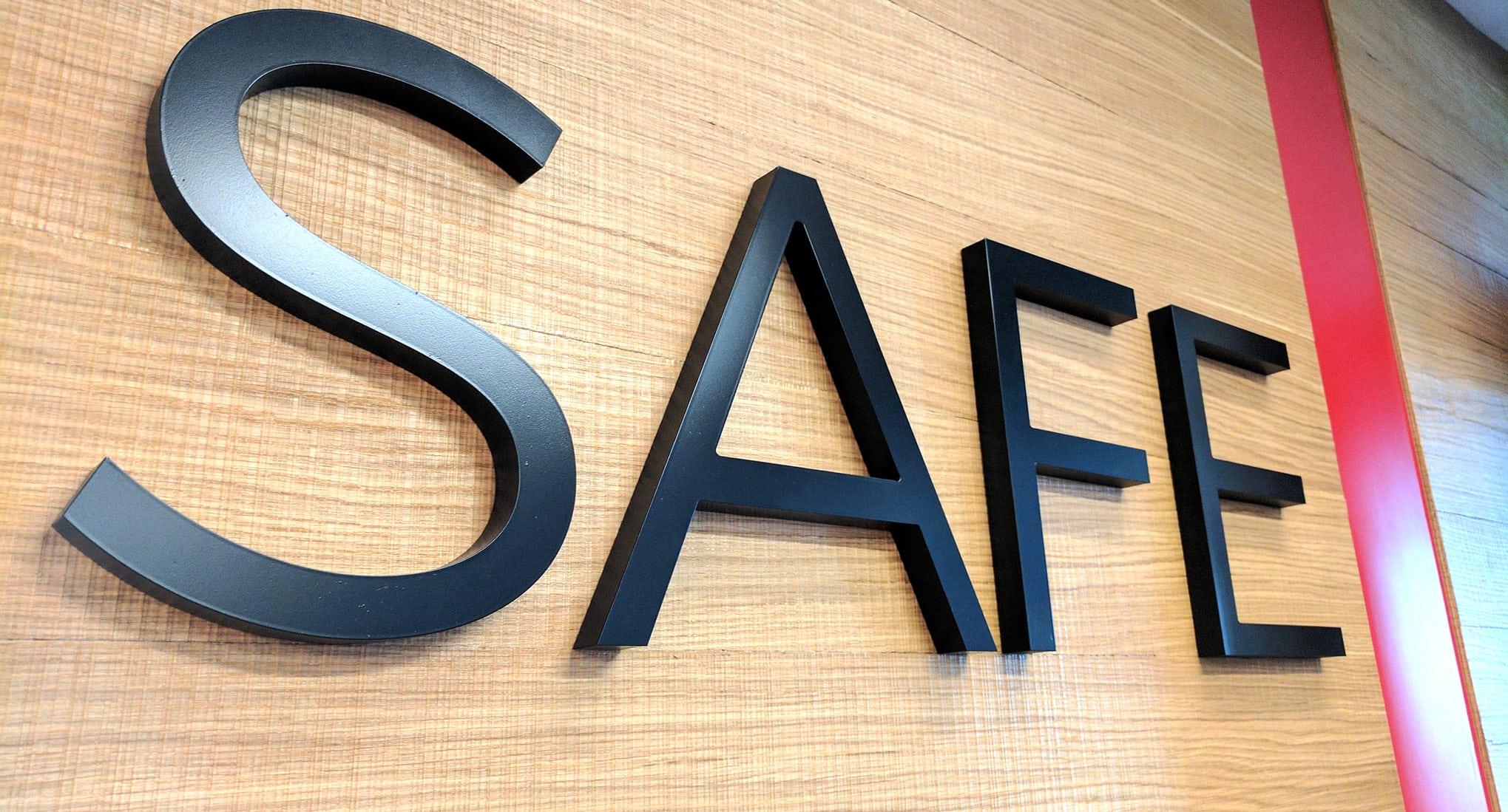 Descripción de la imagen: Una foto del logotipo de SAFE en una pared. La palabra &quot;SAFE&quot; está en letras negras junto a una línea roja a la derecha. La pared es de madera rubia clara.