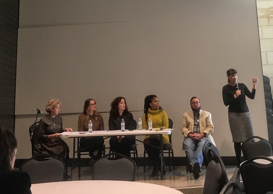 A panel of four female-identified people sit at a table along with one male-identified person. A female identified person and standing, holding a microphone.