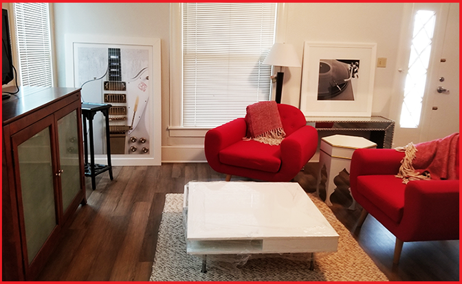 A beautifully furnished home space with a white coffee table in surrounded on top of a rug. Surrounding the table is a red chair, a red sofa, a TV stand, and a TV. The items are all on a hardwood floor.