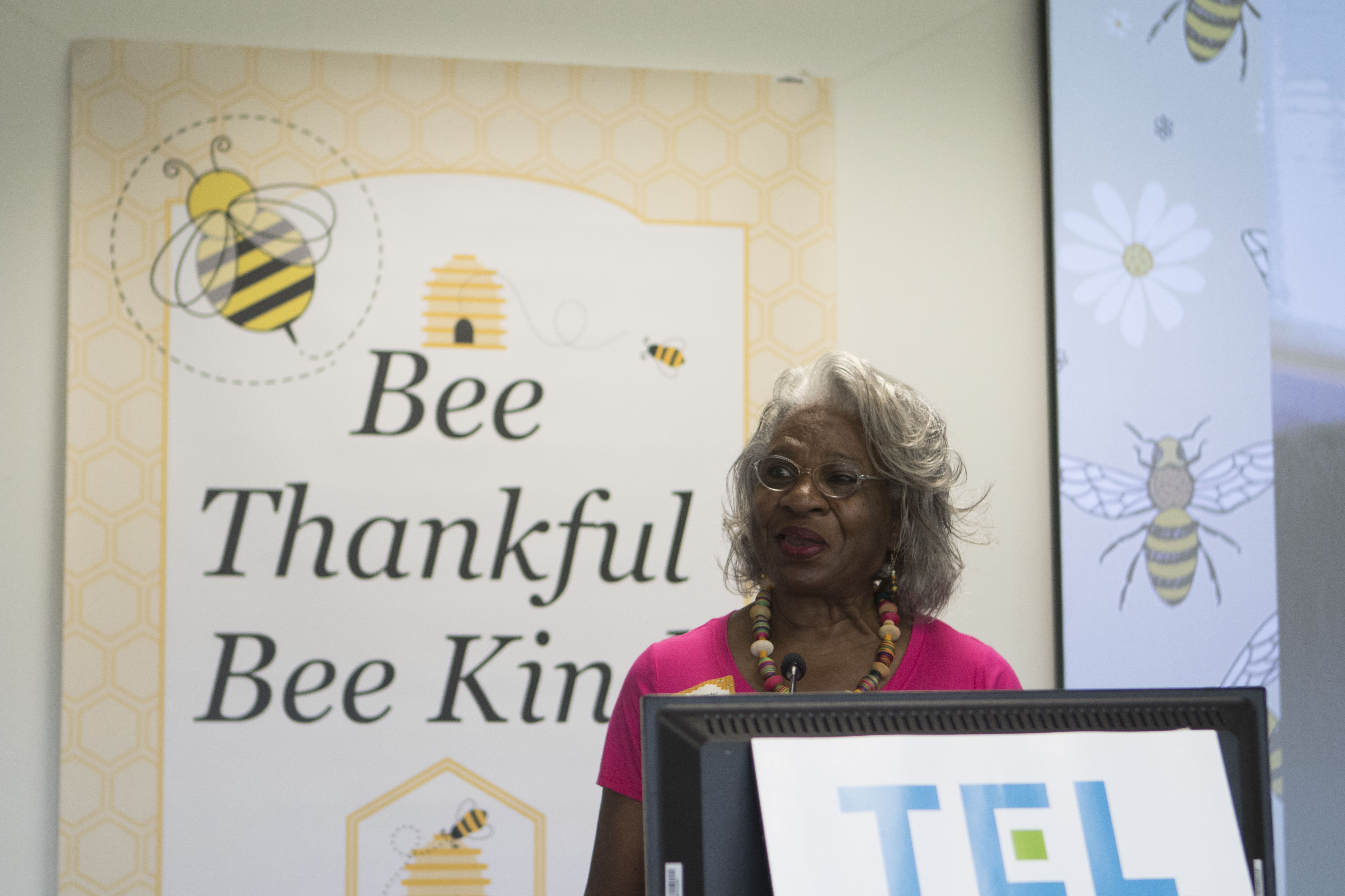 A female-identified person stands at a podium delivering a speech. A sign behind her reads "Bee Thankful Bee Kind"