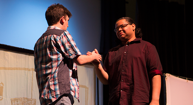 Two male identified people are on stage during a live play. They are shaking hands. One&#039;s back is facing away from the camera and the other is facing the camera.