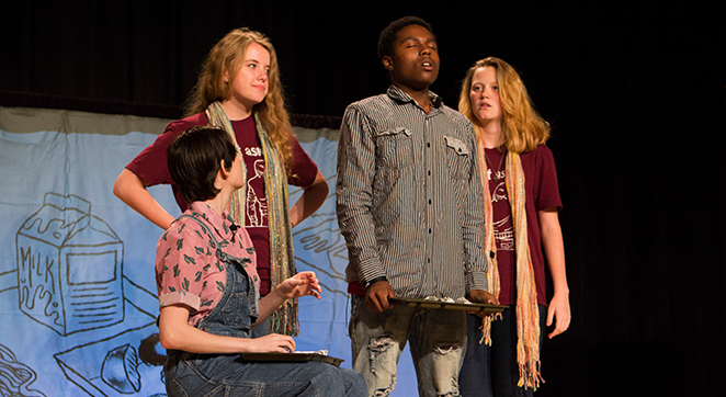 Four teens are together on a stage. Three are standing, one is sitting in a chair.