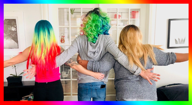 A photo of three people with their back to the camera. Two people have rainbow-dyed hair. They are standing triumphantly with their arms around each other.