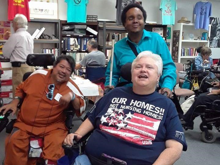 A photo primarily featuring Nancy Crowthers, who is sitting in a wheelchair with a smile on her face. Two people are behind her, one standing, one in a wheelchair. They are also smiling.