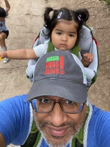 Una selfie de Pranish Kantesaria. Un niño pequeño está detrás de Pranish. Pranish lleva un sombrero de Austin Pets Alive y sonríe a la cámara.