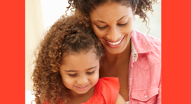 A photo of a woman smiling. She is standing close to her young daughter who is also smiling, though not quite as expressively as her mother.