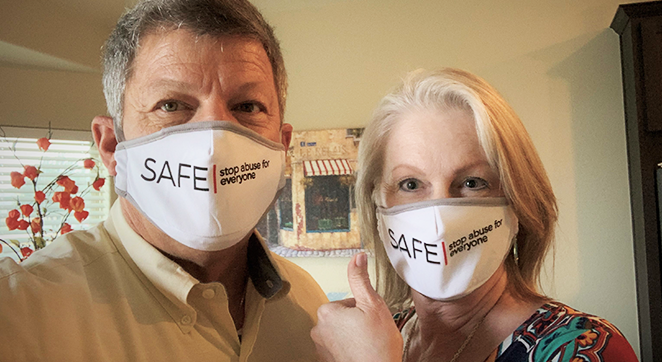 A photo of two people standing next to each other, facing forward. They are wearing masks that say SAFE: stop abuse for everyone on them.