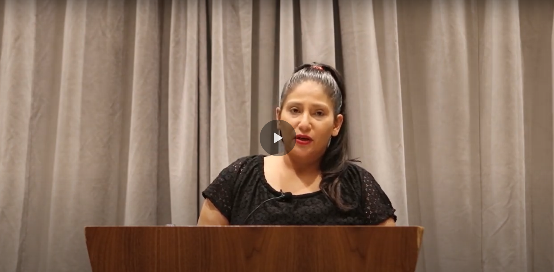 A still image from a video. The image shows a woman standing at a podium giving a speech.