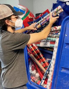 A photo of a person wearing a mask. He is placing rolls of wrapping paper in a large, blue bin.