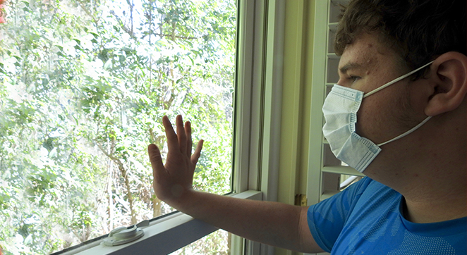 A photo of a person wearing a mask. The person is touching a window wit hone hand. Pale green trees are visible through the glass window. The person is wearing a blue shirt.