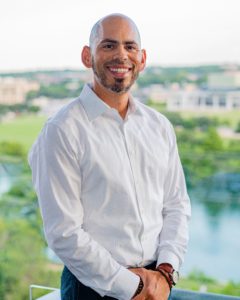 A photo of a man named Greg Galindo. The man is facing the camera wearing a white, button up shirt. He has a trim beard. There is greenery, water, and buildings in the background.