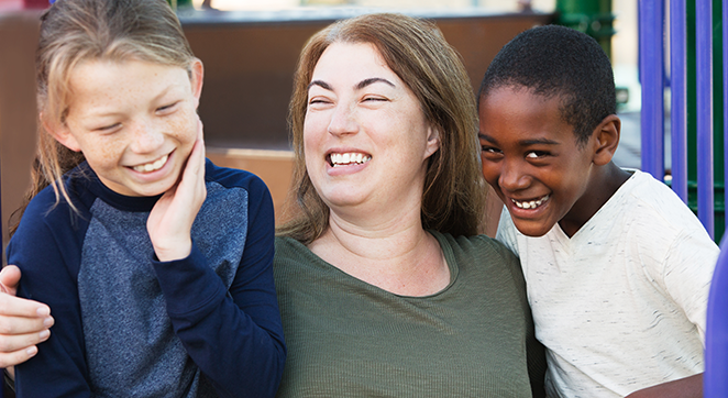 Image description: A stock photo depicting foster parenting. A white woman is sitting down with her arms around two children. They are all smiling. One child is to be a young, Black boy and the other child is a young, white girl.