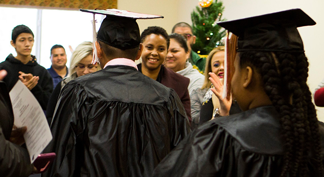 Image description: A photo of the backs of several students dressed in black caps and gowns as during a graduation ceremony. Family and friends are smiling in the background.