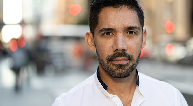 Image description: A stock photo of a Latinx/Hispanic man looking at the camera with a serious expression. His hair is dark and he has a short beard. He is wearing a white shirt. An out-of-focus city environment is behind him.