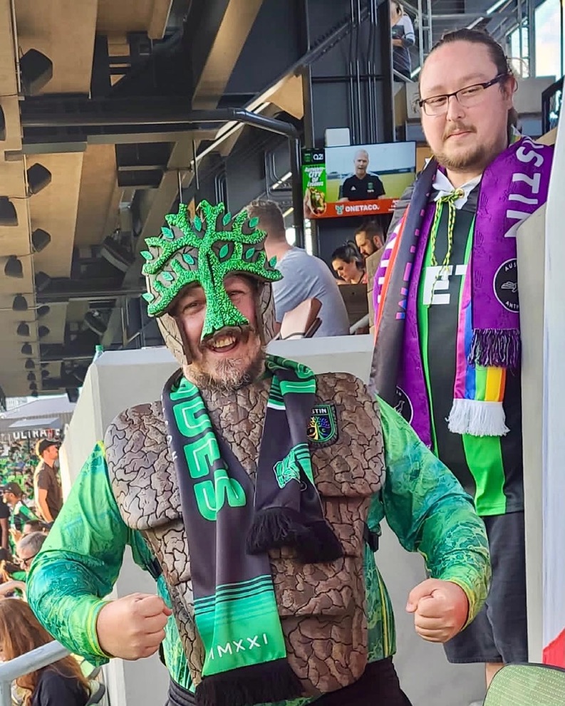 Image description: A photo of two male-presenting people at the first home game of the Austin FC. One person is wearing a Pride rainbow scarf. The other person is wearing a mask in the shape of a tree, which is the Austin FC logo.