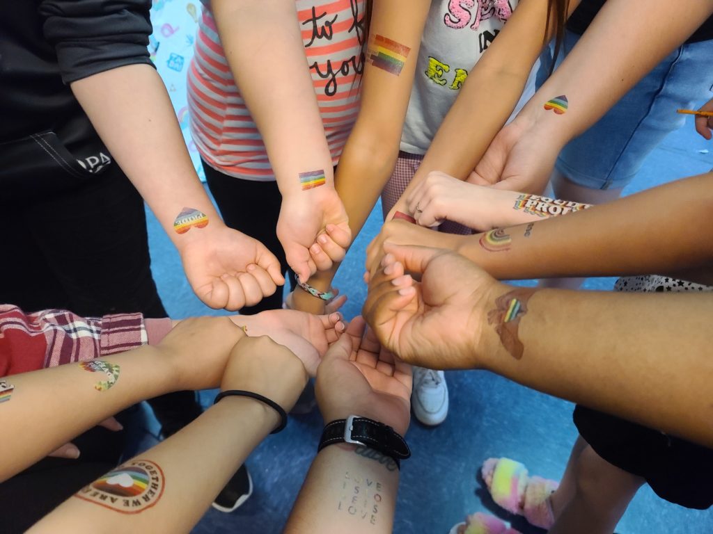 Descripción de la imagen: Los niños de SAFE que llevan tatuajes temporales en los brazos se reúnen en círculo para mostrar sus tatuajes del Orgullo. La foto sólo muestra sus brazos con las manos juntas en el centro. La mayoría de los tatuajes muestran arco iris en diversas formas, como corazones, banderas.