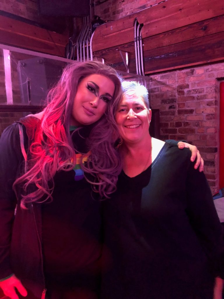 Descripción de la imagen: Una foto de dos personas en un bar/club. Una de las personas va vestida de travesti, lleva el pelo largo y rosa y un maquillaje precioso, con unas alas grandes y preciosas y una sombra de ojos que complementa su pelo. Están junto a su madre, que sonríe cariñosamente a la cámara.