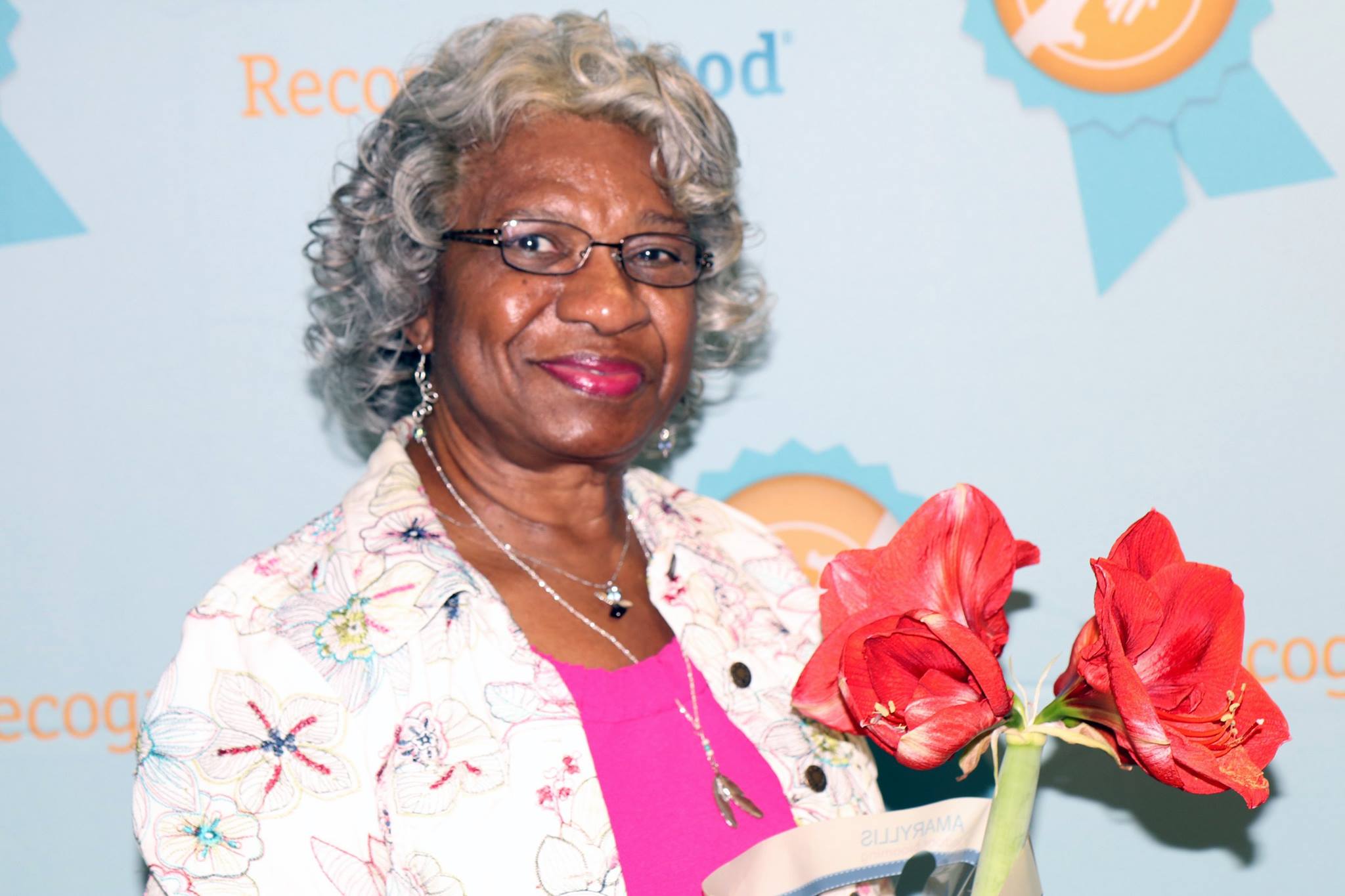 Image description: A photo of Frankie Fowler smiling at the camera. Frankie is a Black Woman with grey hair and dark-framed glasses. She is wearing a white top with a lovely flower pattern on it. Beautiful red flowers are in the foreground.