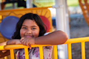 Image description: A stock photo of a young Latinx girl. She has dark hair and is smiling at the camera. She is leaning on a yellow playground fense.