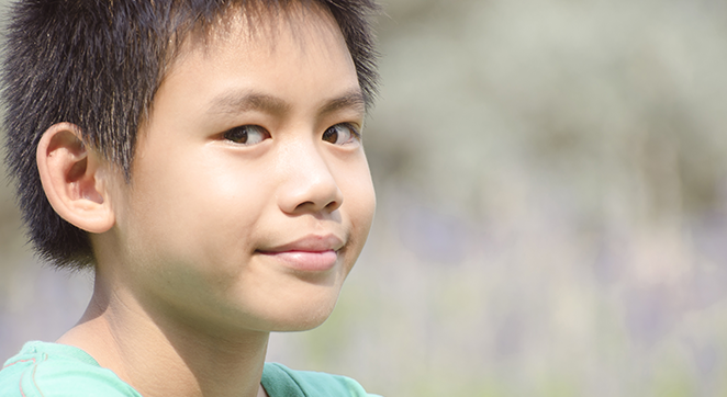 Image description: A photo of a young, Asian boy looking at the camera. He has dark hair and a very faint smile. The background is a tan outdoors setting that is blurred by the depth of field.