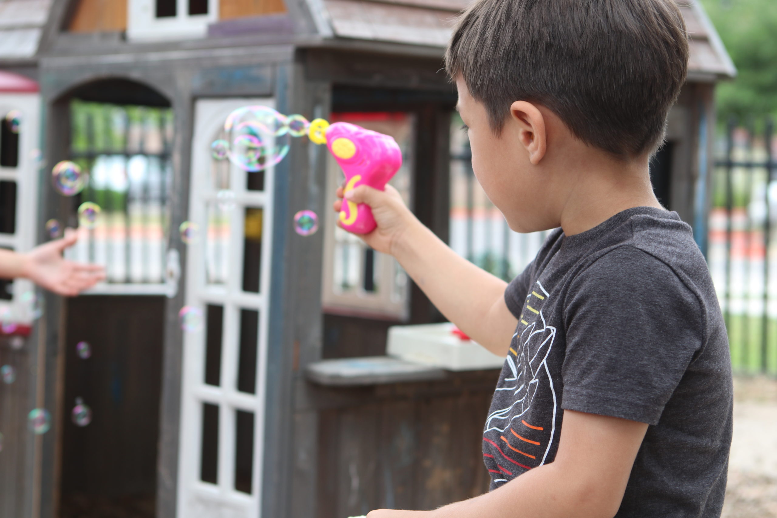 Image description: A photo of a young boy holding a a pink bubble gun. The gun is producing little bubbles. The boy's back is to the camera.