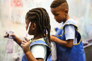 Image description: A photo of two young children wearing smocks. They are playing at an art exhibit at Thinkery.