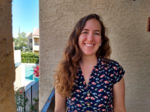 Image description: A photo of a woman with brown hair smiling at the camera. She is wearing a colorful top. A stucco wall is behind her. 