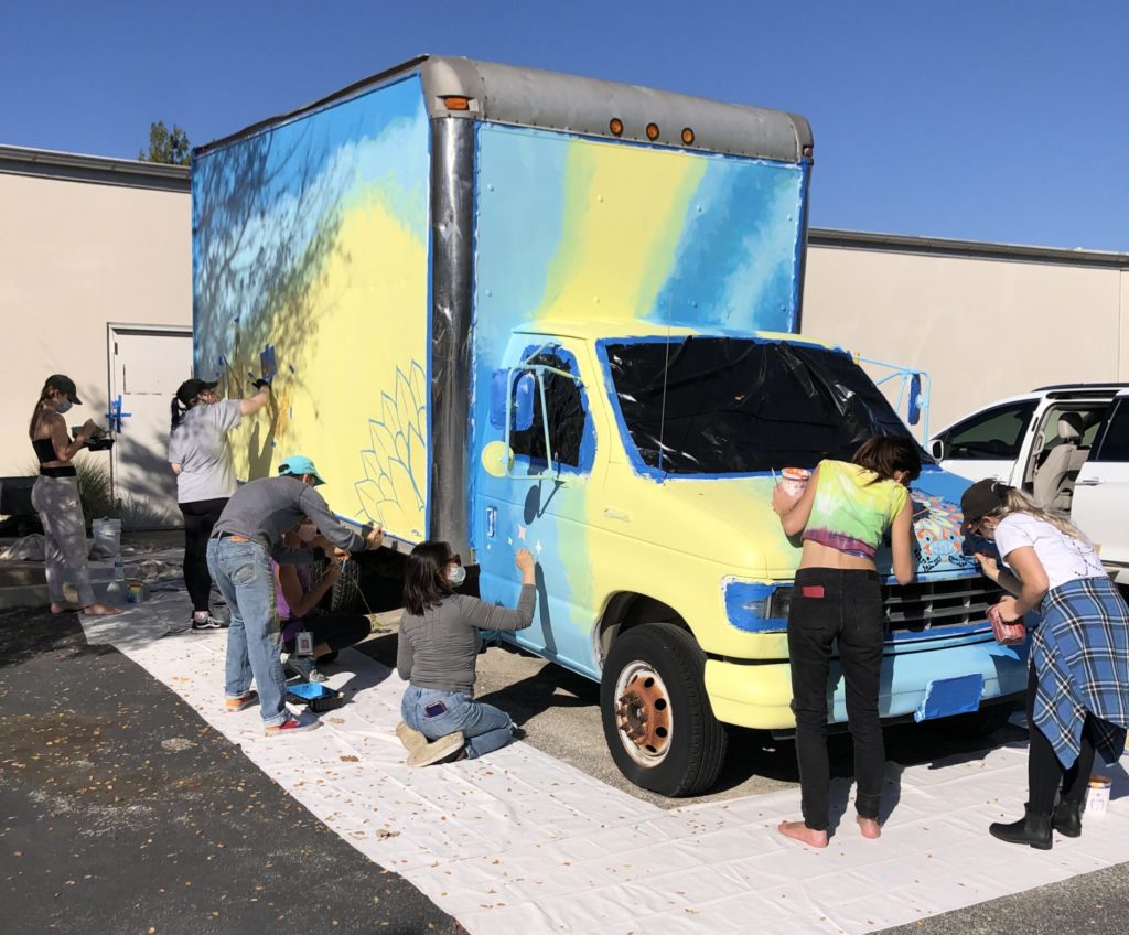 Image description: A partially painted truck. Bright blues and yellows cover the truck. Volunteers are surrounding it while they paint.