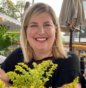 Image description: A photo of a white woman with blond hair. She is smiling and facing the camera while wearing a black blouse. A green plant is in the foreground and a closed parasol is in the background.