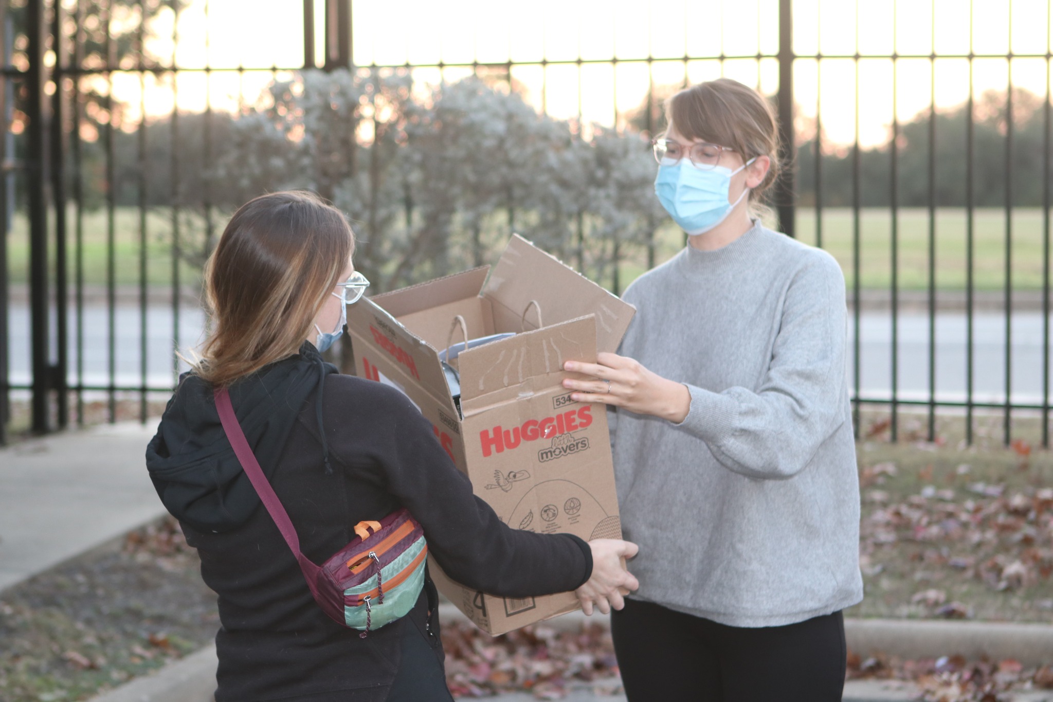 Image description: A photo of a person in a grey sweater accepting a box of gifts from another person who is wearing a black jacket and cute purse. They are both wearing masks and standing in the SAFE Family Shelter parking log.