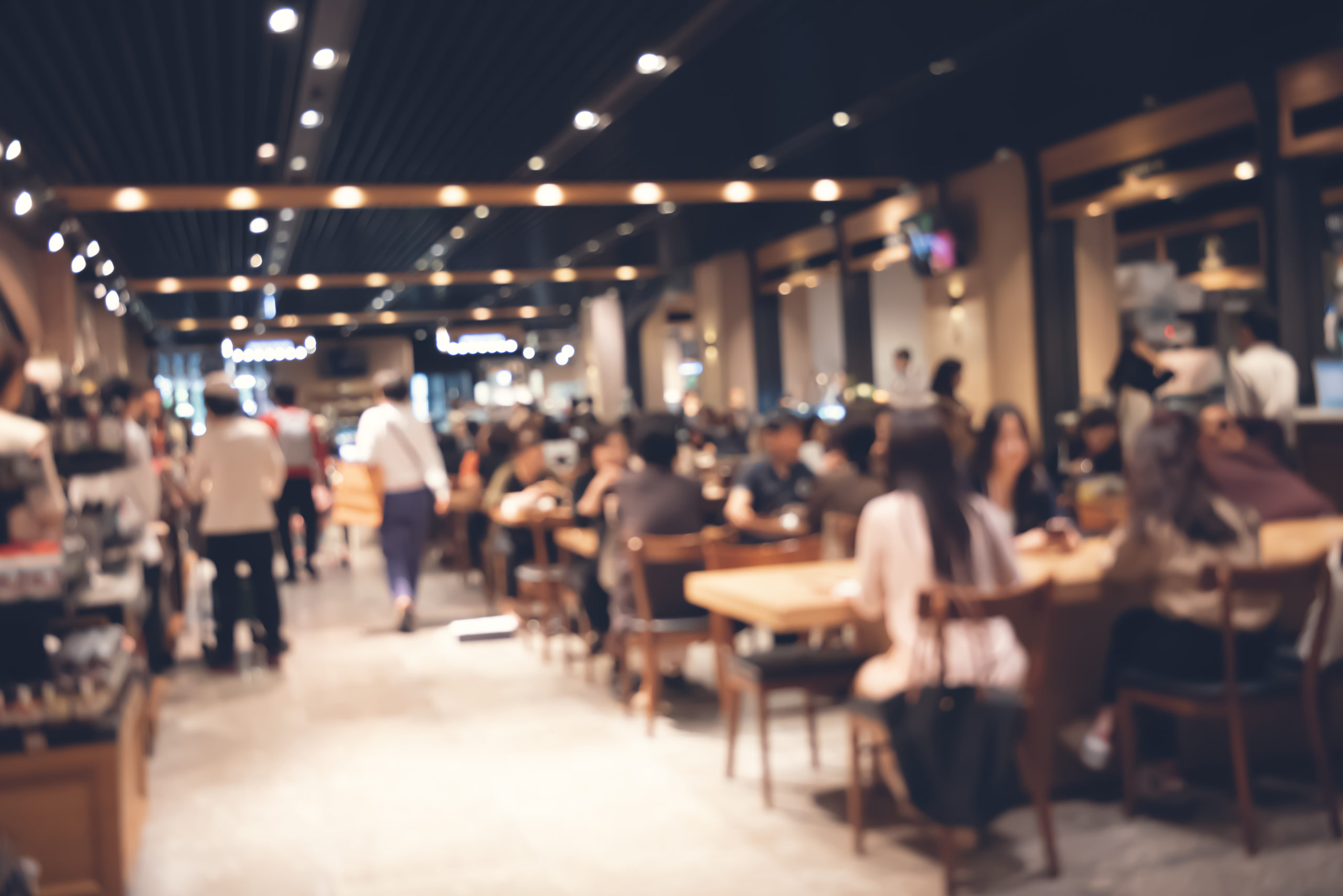 Image description: A stock photo of a cafe with many people sitting at tables and walking around. The image is intentionally blurry.