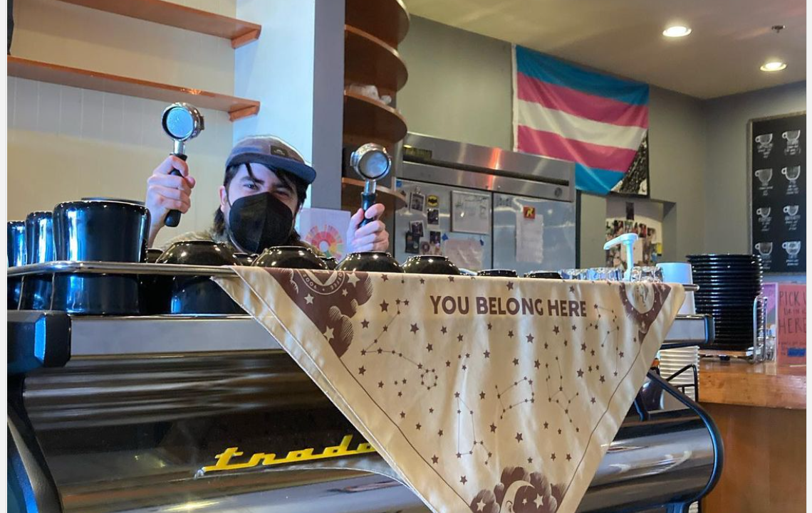 Descripción de la imagen: Un barista de pie detrás de una barra sosteniendo dos portavasos de cafetera. Una bandera trans está en el fondo y una pancarta que dice &quot;you belong here&quot; está delante de la barra.