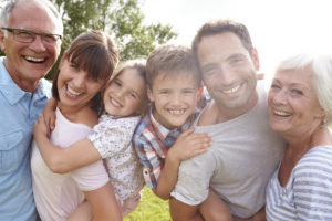 Descripción de la imagen: Foto de archivo de una familia blanca multigeneracional. Todos miran a la cámara sonriendo. Están al aire libre en un buen día.