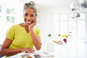 Descripción de la imagen: Una mujer negra/afroamericana sentada en una mesa sonriendo. Su cabeza está apoyada en su mano. Está mirando una revista sentada en una mesa blanca e impecable.