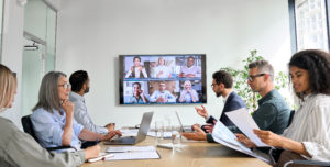Descripción de la imagen: Foto de archivo de un grupo diverso sentado en una mesa de conferencias en el lugar de trabajo. Varias personas están en persona y varias están en una pantalla de televisión, asistiendo a distancia.