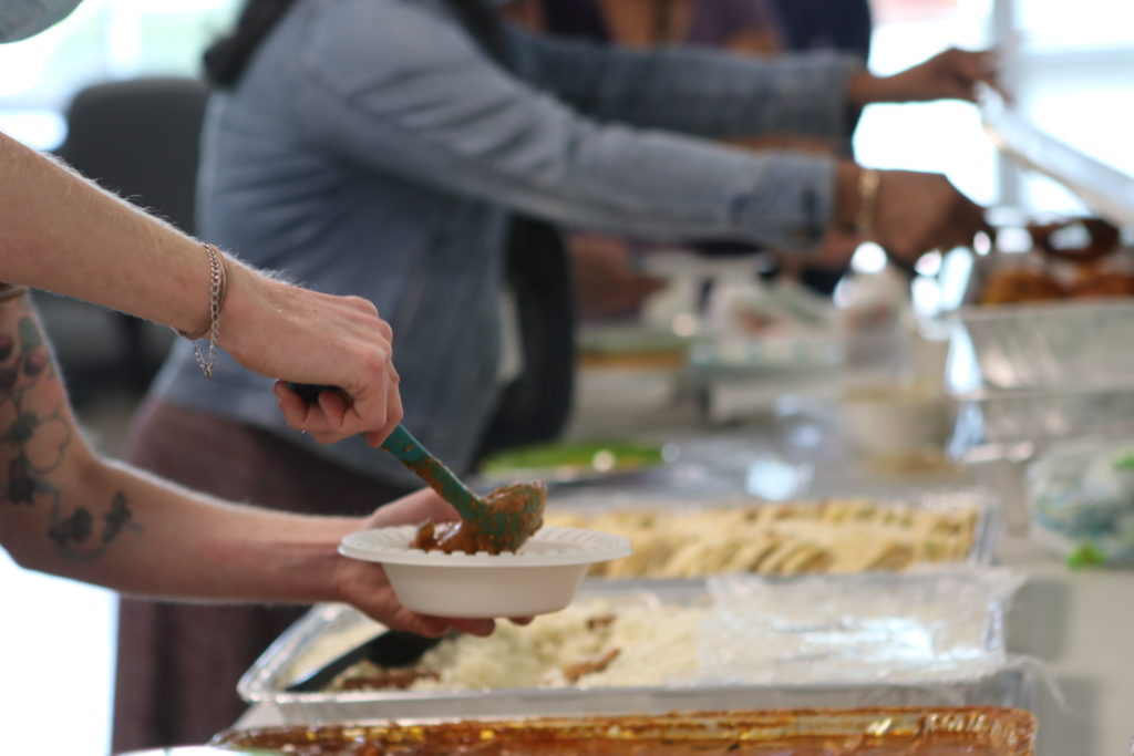Image description: A photo of hands putting pouring food into a bowl and reaching for food during SAFE's Asian, Pacific Islander, and Pan Asian potluck.