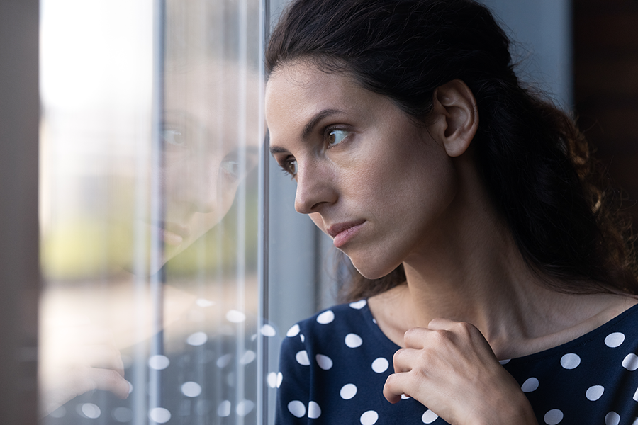 Image description: A stock photo of a woman looking forlorn out a window. She is wearing a dark top with white polka dots.