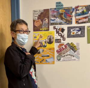 Image description: A photo of a woman wearing glasses and a mask pointing at collages she made on a wall.