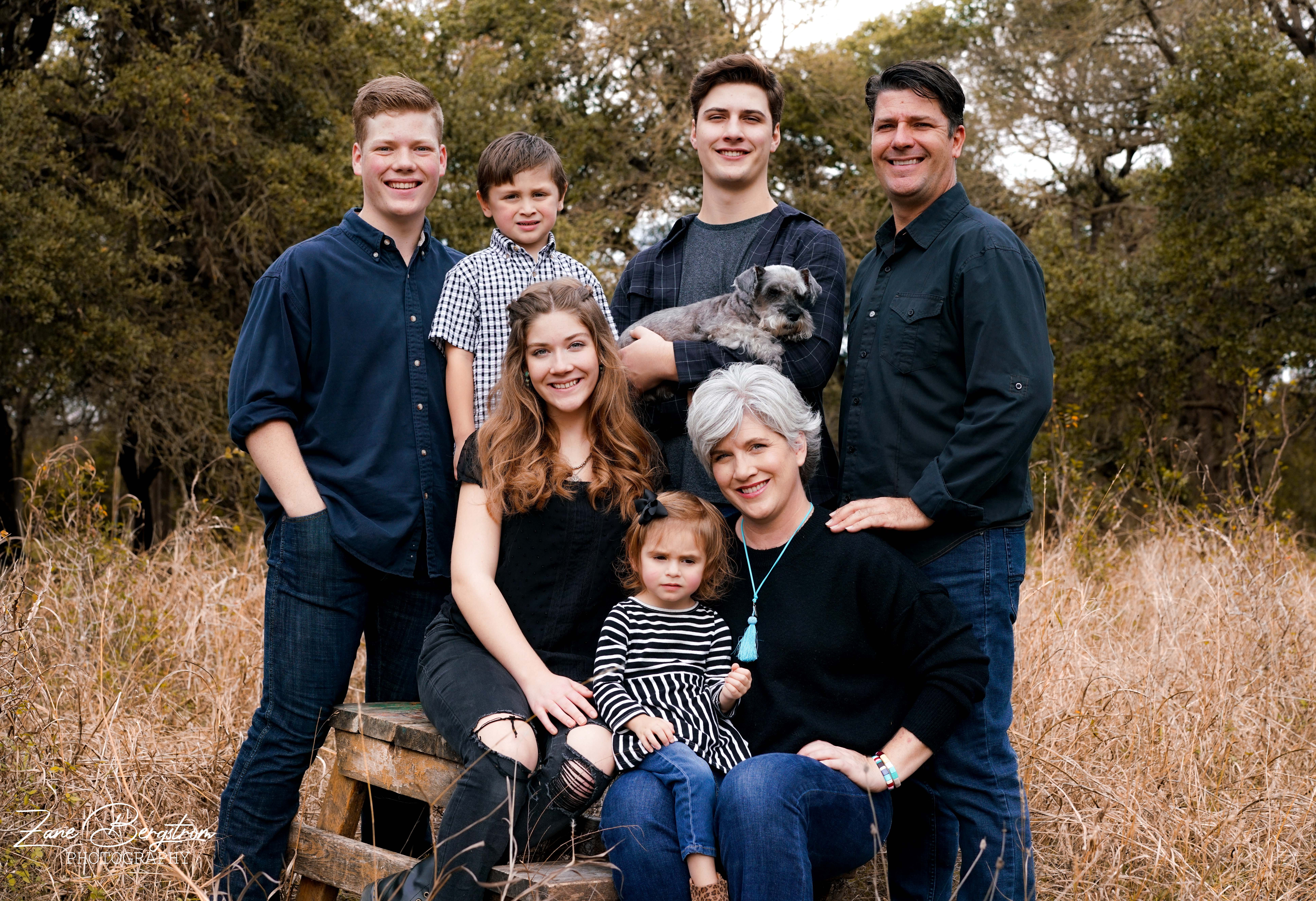 Image description: A family photo. Two adults and their five children are together outside smiling at the camera.