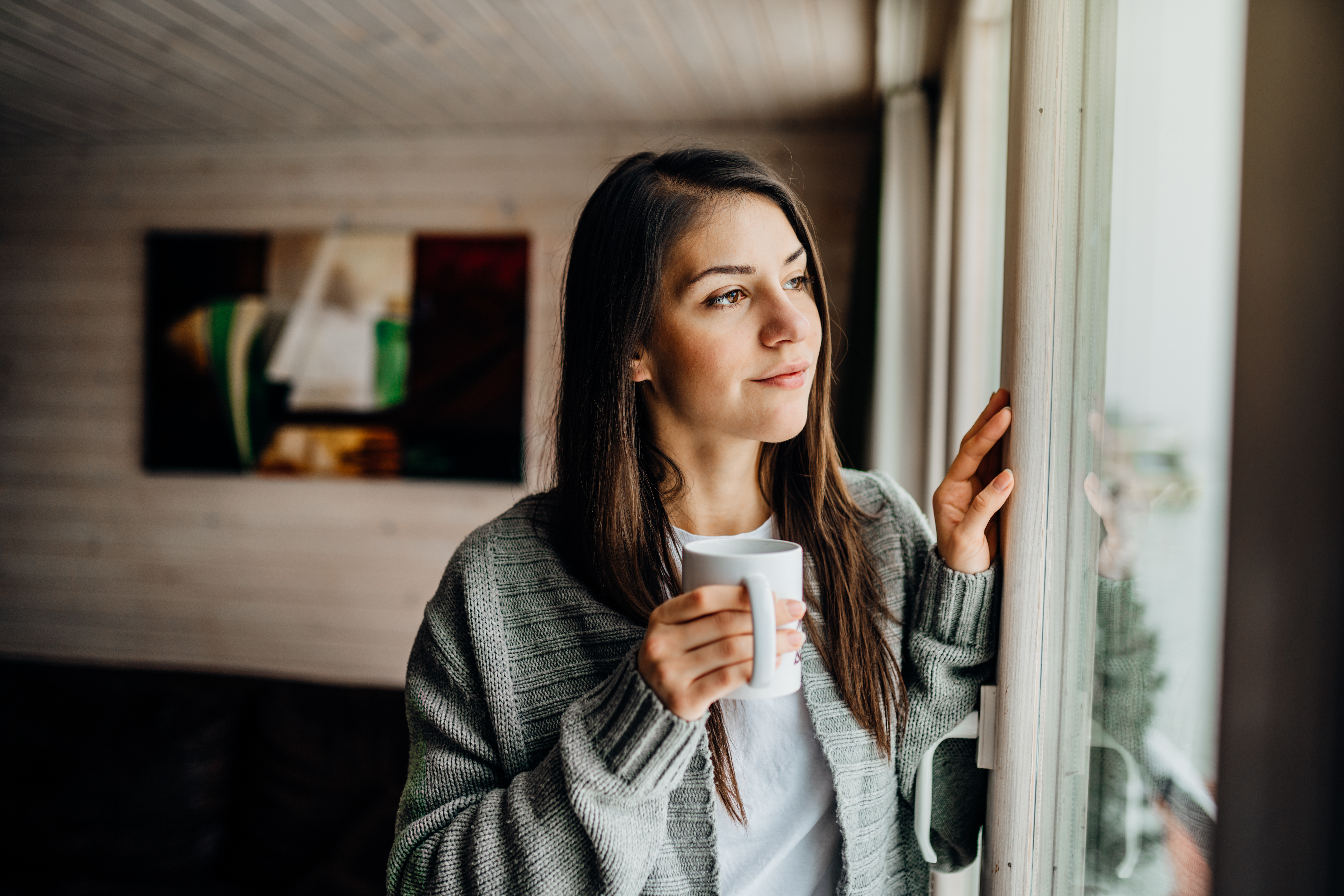 Young woman holding coffee looking out home window.